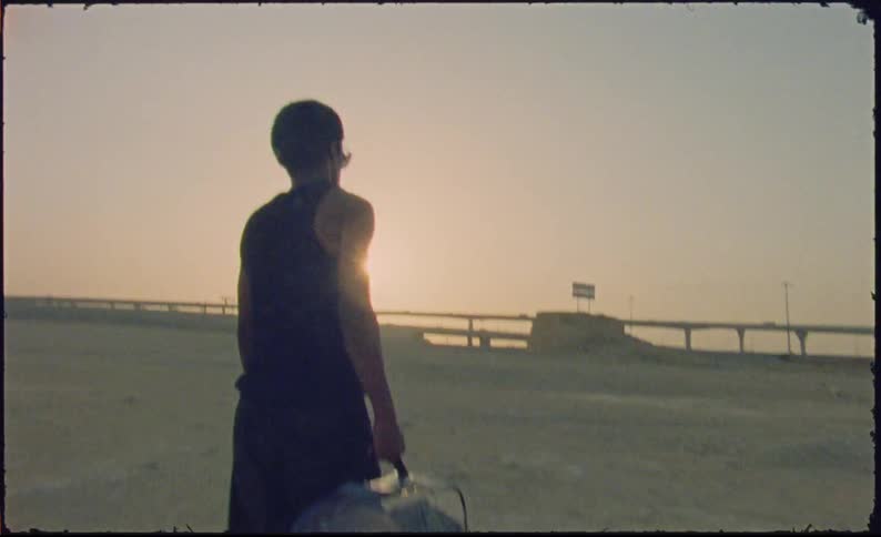 A young man carries a transparent suitcase walking through a barren terrain near a highway at sunset
