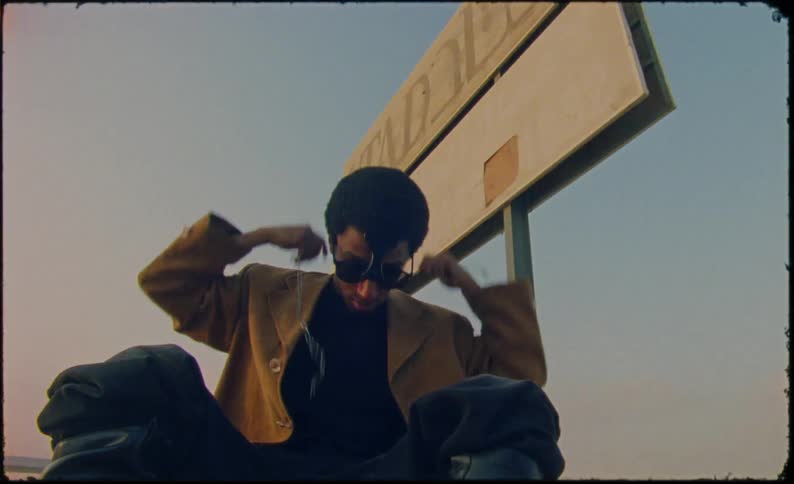 Man wearing a fashionable outfit wraps a chain around his neck while sitting on the ground near a sign