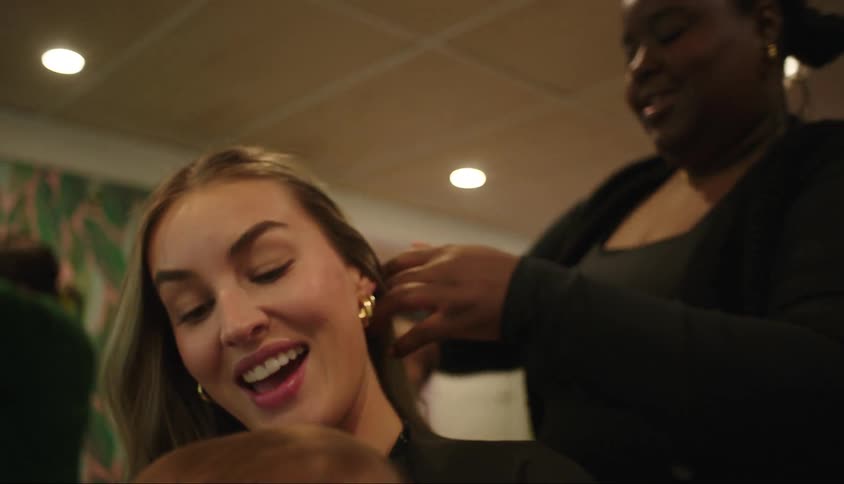 A woman with a baby on her lap sits in a salon chair while a stylist combs and styles her hair
