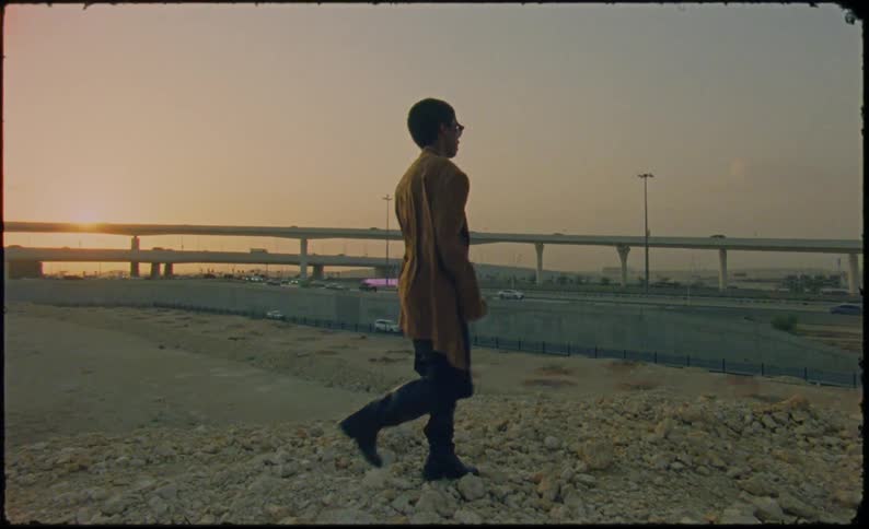 Young man wearing a long brown coat while standing on a rocky embankment in front of a highway