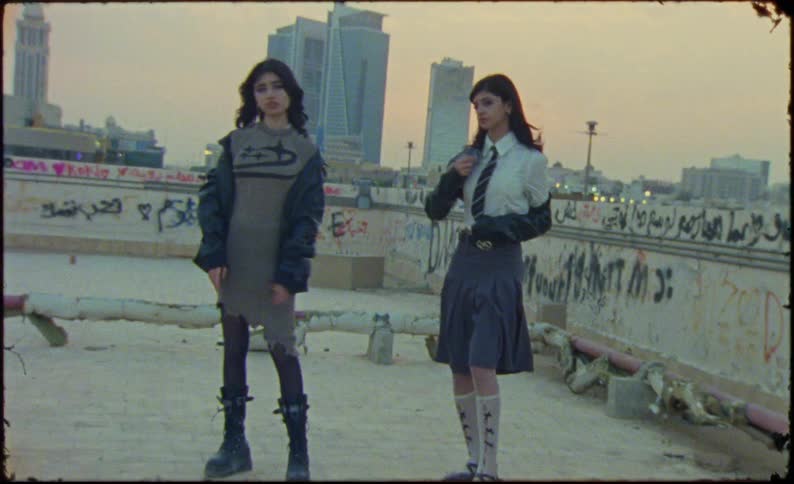 Two women model outfits while standing near a graffiti covered low wall on a building roof after dusk