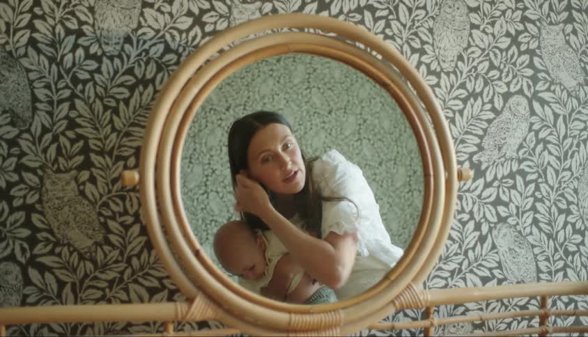 A woman holding a baby on her lap and is applying makeup to her face looking into a wooden framed mirror