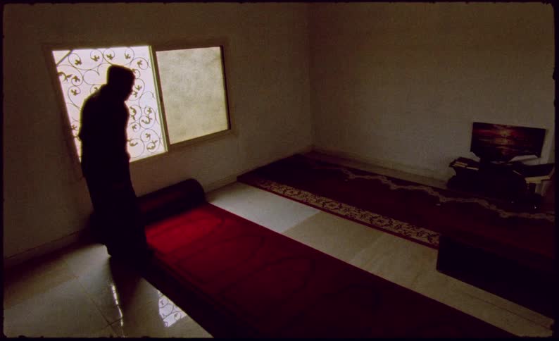 Silhouette of a robed figure praying on a room with carpets and sparce furniture