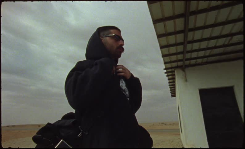 Man with sunglasses and sweatshirt next to a house taking off his backpack