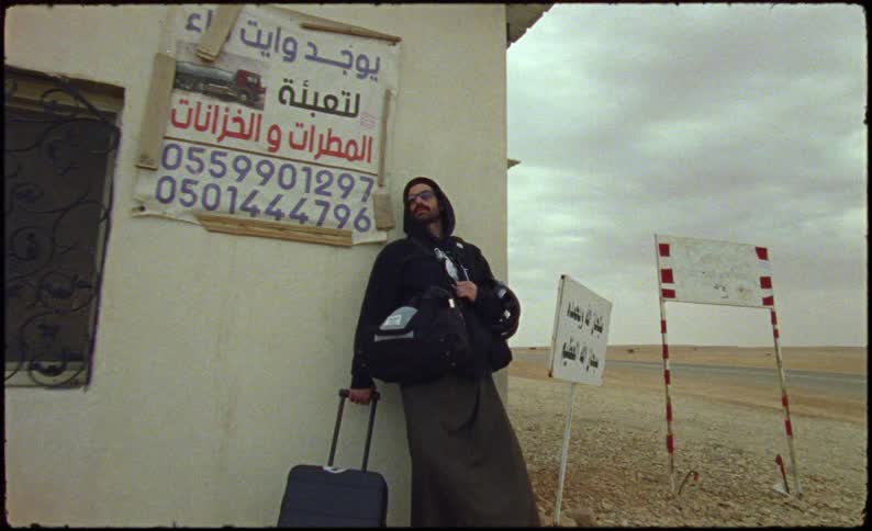 Man with sunglasses and a black top holding a suitcase leaning against a wall on the side of a road surrounded by a desert environment
