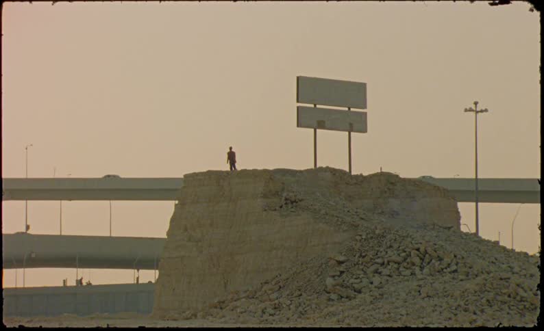 Man standing on a hill of rocks next to an overpass with vehicle traffic in the late afternoon