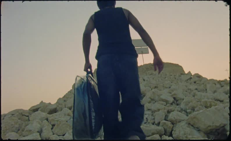 Man carrying a bag walks up a mound of rocks toward a sign under a red tinted sky