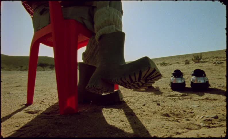A person sitting on a red plastic chair wearing black boots in a desert region