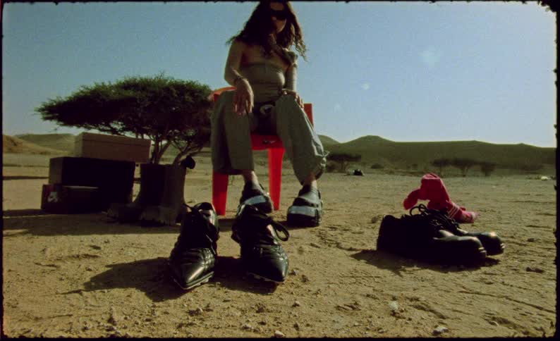 A young woman sits on a plastic chair in the desert experimenting with various types of shoes