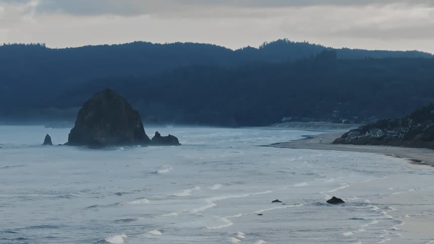 Waves pass rock formations before rolling onto beach near forested mountains on a foggy overcast day