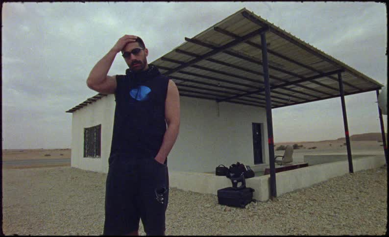 Man in shorts and tank top sweatshirt running his hand through his hair standing outdoors next to a house in a deserted environment