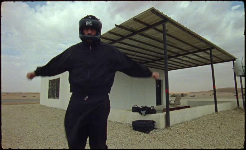 A man standing in black clothes and a black helmet is stretching his arms and behind him there is a house in a rural region