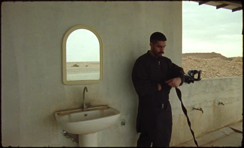 The man dressed in all black is wrapping his hand with a black wraphe is standing beside of a mirror and a white pedastal sinkthere are two water spigots to the right of him