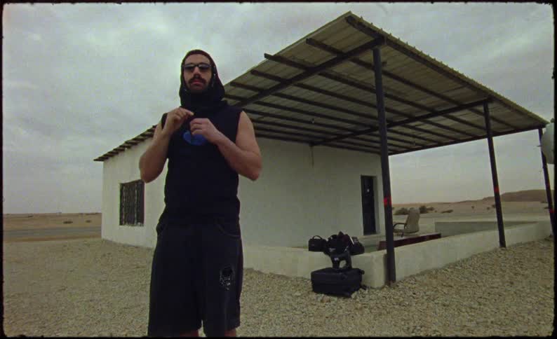 Man next to a house in a desert environment pulling on the hood of a tank top