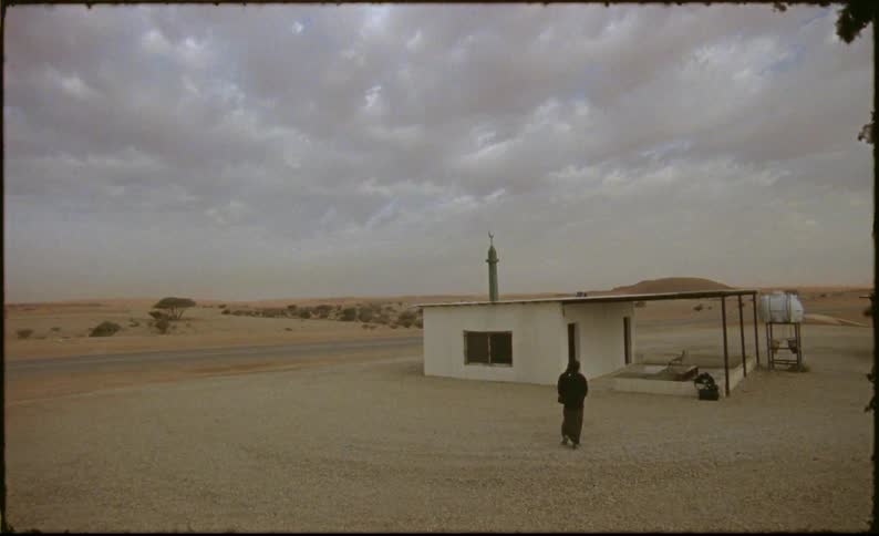 A person wearing black walks around a solitary building with a flat roof in a vast arid landscape