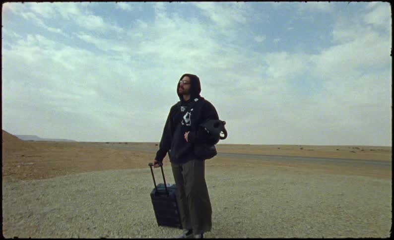 Man stands in the gravel with a suitcase near the road in a desert on a sunny day