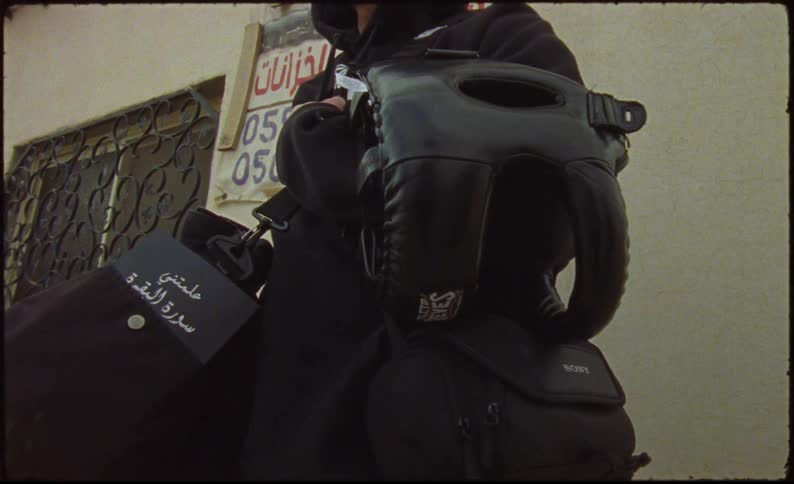 Man carrying a backpack and a book stands near a building with an arabic sign