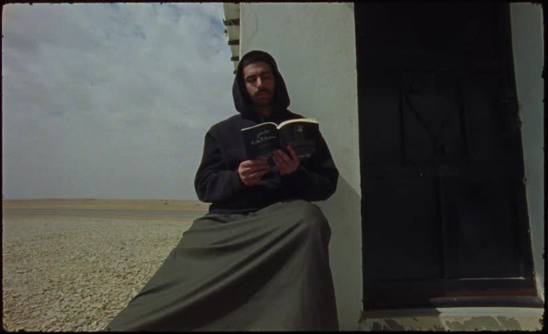 Man sitting outdoors near the door to his house reads a book while a highway passes through the arid land in the background