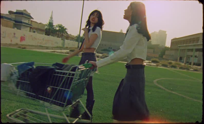Two girls in school clothes are walking on a football field lawn carrying a grocery cart