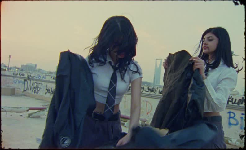 Two girls are on a roof top and are taking clothing out of a shopping cart to hold up in front of themthey then spin and look at the camera
