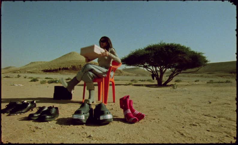 Woman sitting on an orange chair in a desert environment and opening a shoe box