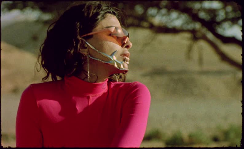 Model woman wearing a long pink dress sitting on an orange chair with shoes around her in a desert environment