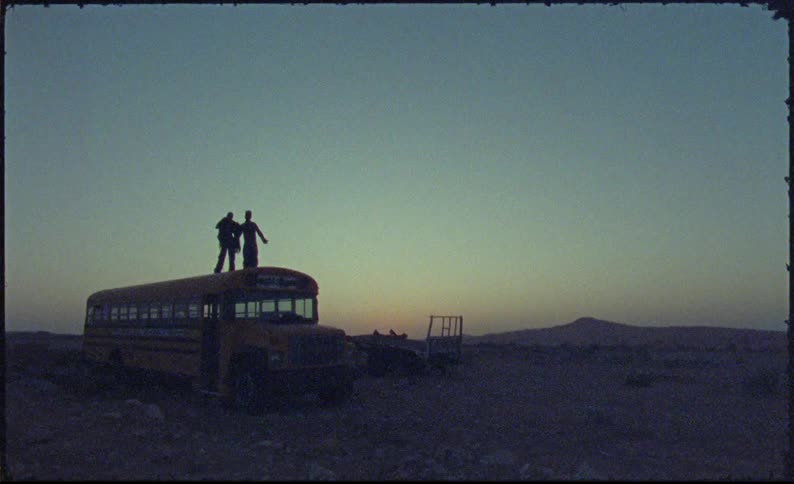 Two dancers dance on top of a school bus parked in the desert after dusk