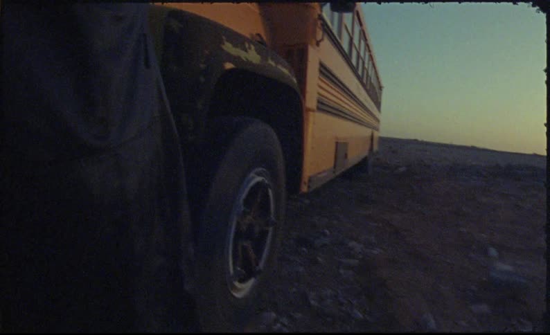 A person wearing a headscarf and a coat climbs over a yellow schoolbus parked in the desert