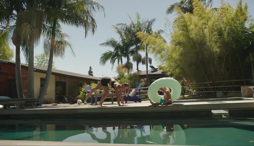 A person performs a handstand by a pool while another person sits nearby with a large pool float and palm trees in the background