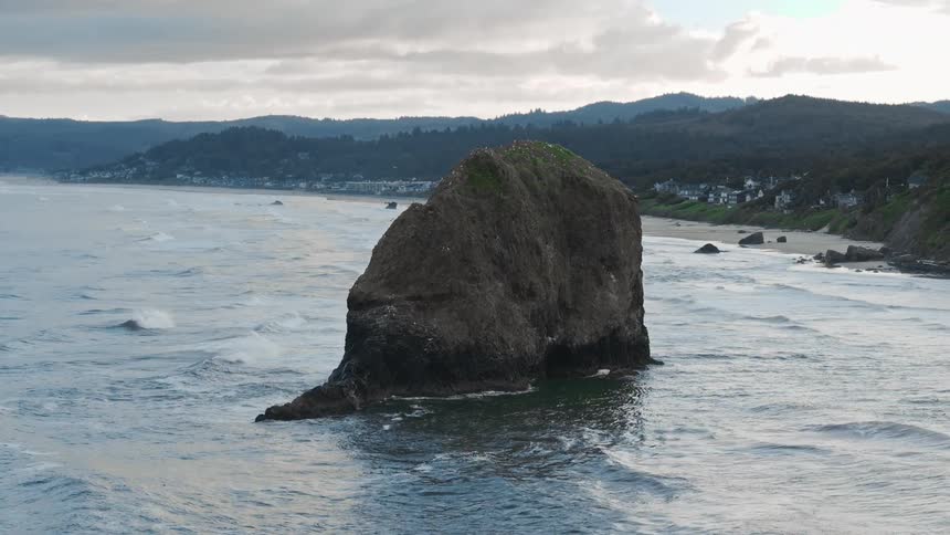 A view of large rock formations in the ocean near a coastal town with a backdrop of misty mountains and cloudy skies