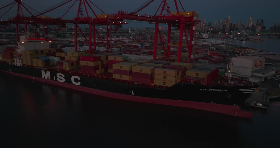 A large barge filled with shipping containers sits in a port near a city