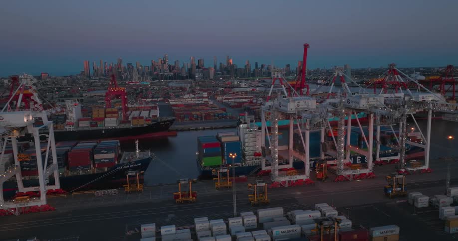 A port with several containers and some orange vehicles passing on a street and it is dusk