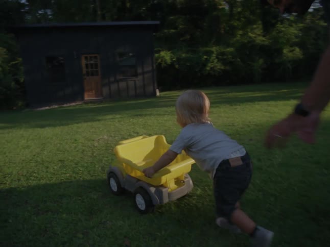 A father follows his toddler son who is pushing a toy truck across a yard