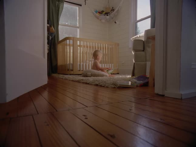 A little baby is on a carpet and playing with a book next to his crib