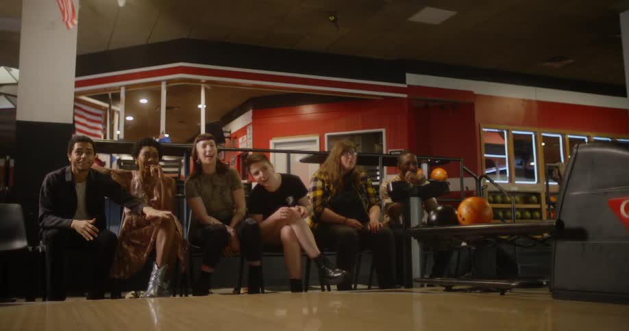 A group of friends in a bowling alley watch as one of them throws the ball down a lane then jump up and celebrate as he comes back and jumps up and rolls on the ground