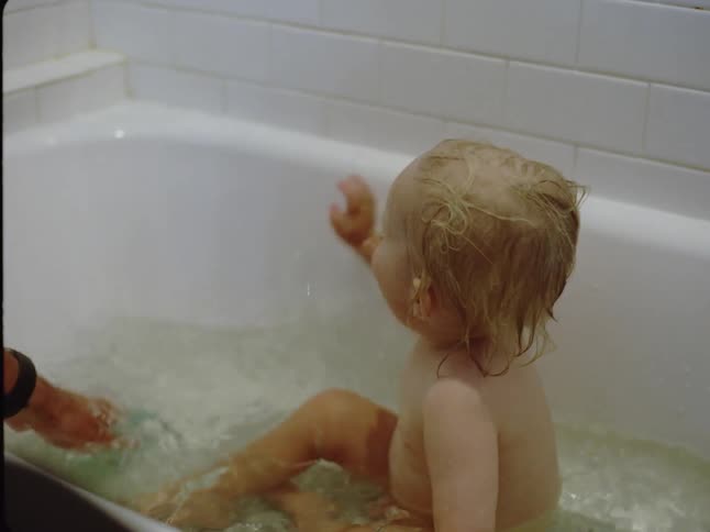 A little blonde baby is in a bathtub taking a bath and is happily splashing his hand in the water