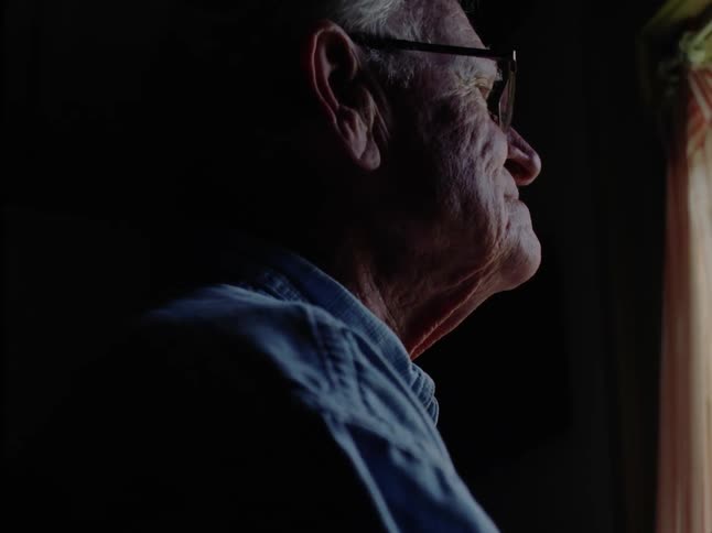 The face of an elderly gentleman with reading glasses and he is in a dark environment looking out of a window