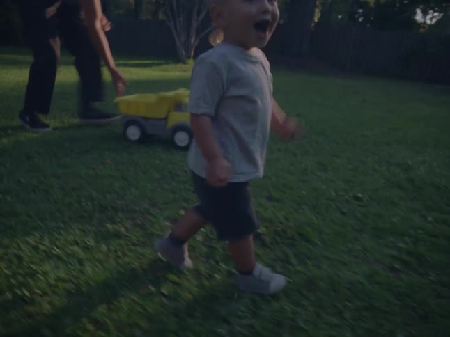 A toddler running on a backyard as his father follows close pushing a toy truck