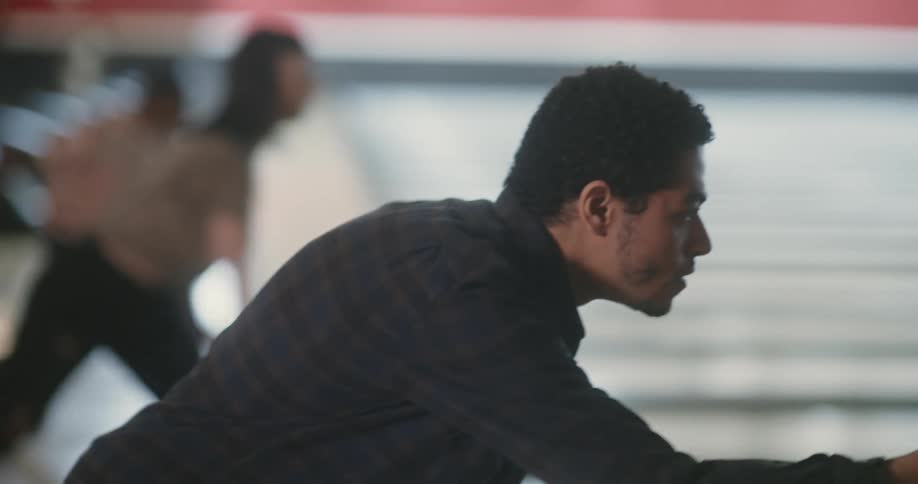 A man in a bowling alley focuses as he throws a ball down the lane as other people in the other lanes do the same then turns around and smiles