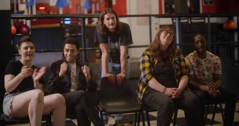 A group of friends sitting in a bowling alley clap and celebrate when another joins them