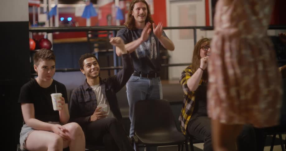 A group of friends cheers in a bowling alley as one of them comes back from a lane and celebrates