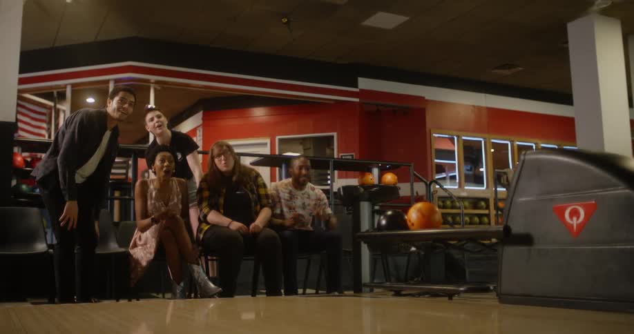 A group of friends in a bowling alley watch as one of them throws the ball then comes back jumping in celebration as they all throw their hands up and slap his