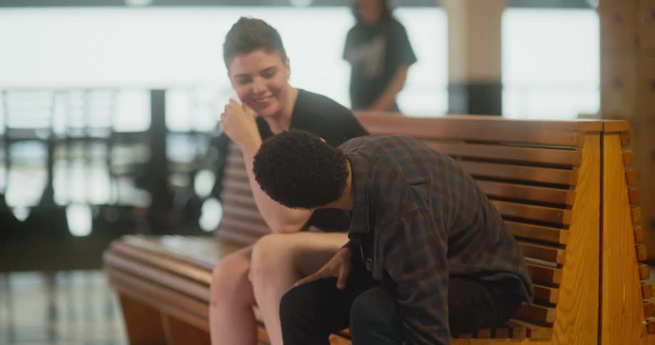 The man is wearing his sneakers and he is sitting next to a woman who is talking to him and then another person arrives and also sits on their bench
