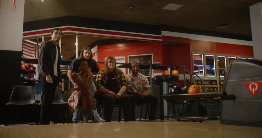 A woman in jean shorts throws the bowling ball and then runs back to the group of four men and one woman and celebrates with high fives