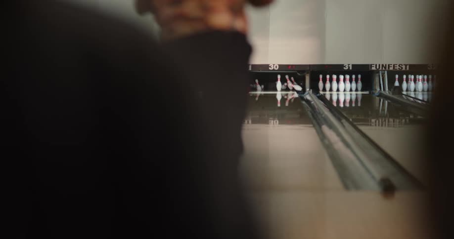 A man in a bowling alley throws a ball down a lane turning back as the ball knocks out all but one pin