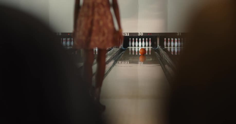 A woman in a bowling alley throws a ball down a lane knocking out all but one pin then turning in place with a half spin