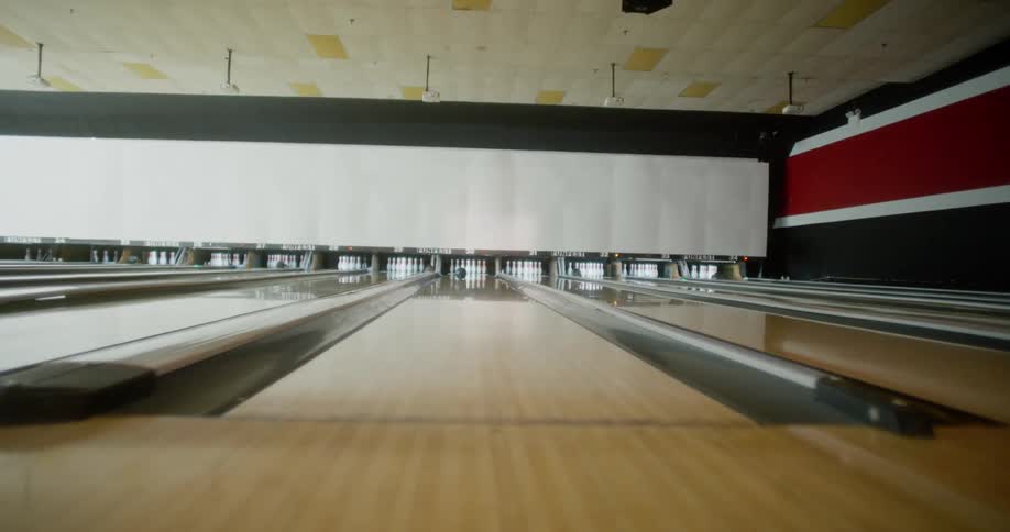 A man in a bowling alley clicks her boot heels together then throws a ball down a lane knocking out three pins