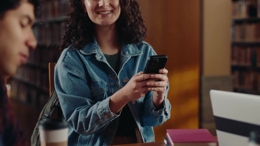 A young woman sitting at a table using her black cell phone in a library