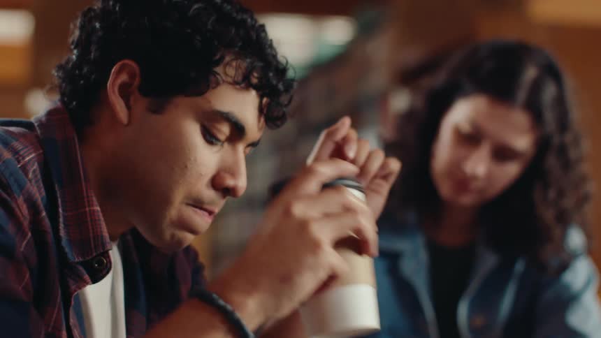 A young man drinks from a cup while sitting next to a woman in a library