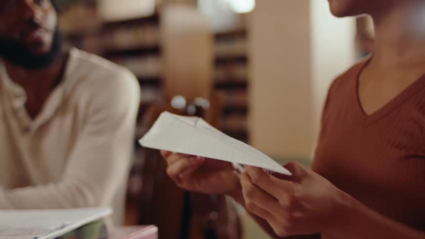 Woman folds a paper airplane while she and another talk at a table in the library with their homework